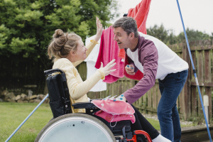Vader en dochter buiten in tuin bij de waslijn. De dochter zit een in rolstoel. Samen hangen ze de was op. Ze hebben er plezier in. 